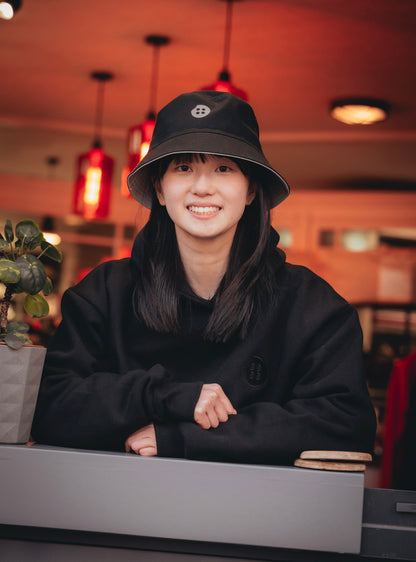 Person wearing a black bucket hat and black hoodie in an indoor setting with warm lighting.